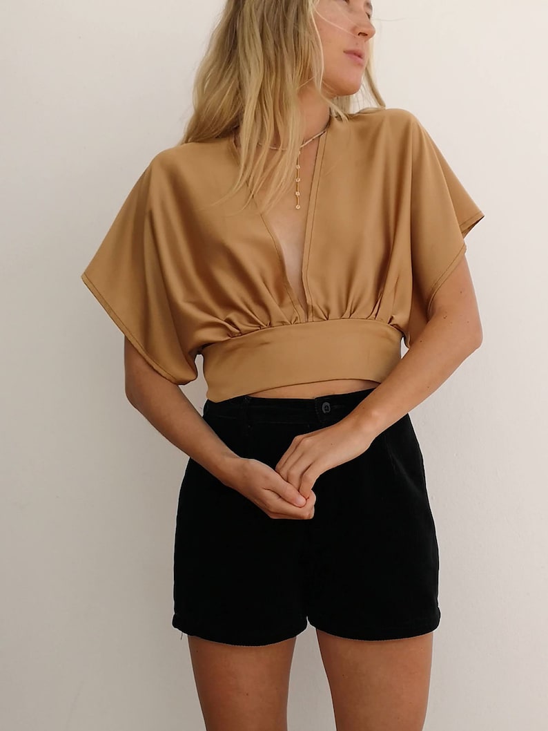 Pu&ograve; includere: Una donna indossa una camicetta di raso marrone con una profonda scollatura a V e maniche corte. Indossa anche pantaloncini neri.