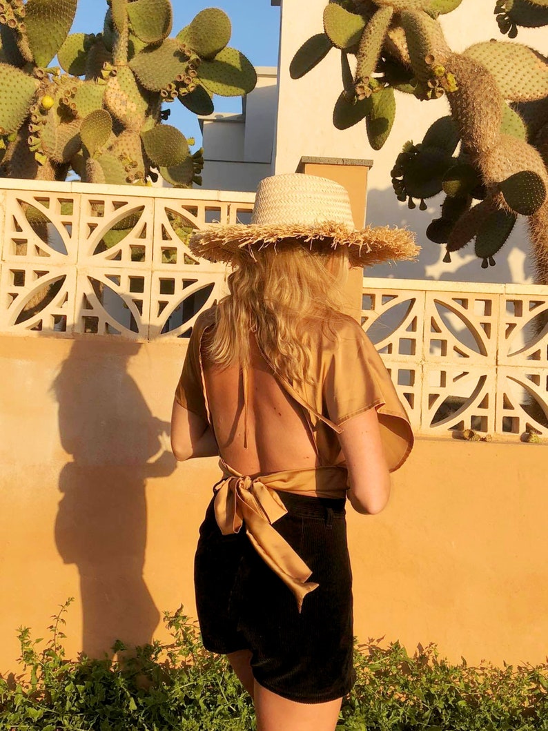 Pu&ograve; includere: Una donna indossa una camicetta marrone chiaro annodata sul retro con un grande fiocco, pantaloncini neri e un cappello di paglia a tesa larga.