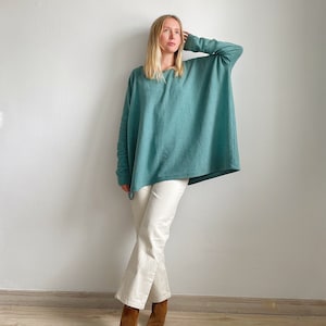 May include: A woman wearing a teal blue oversized sweater with white trousers and brown ankle boots.