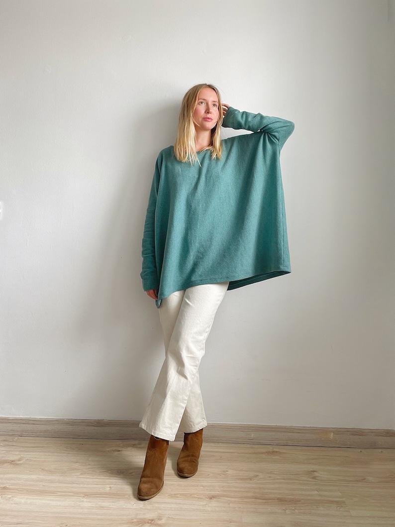 May include: A woman wearing a teal blue oversized sweater with white trousers and brown ankle boots.