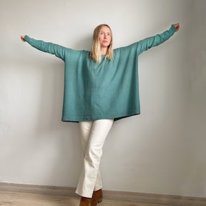 May include: A woman wearing a teal blue oversized sweater with long sleeves and white trousers. She is standing with her arms outstretched and looking up.