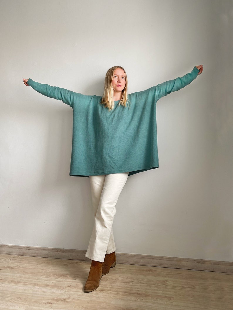 May include: A woman wearing a teal blue oversized sweater with long sleeves and white trousers. She is standing with her arms outstretched and looking up.