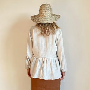 May include: A woman wearing a white, long-sleeved blouse with a gathered waist and a brown skirt. She is wearing a wide-brimmed straw hat.