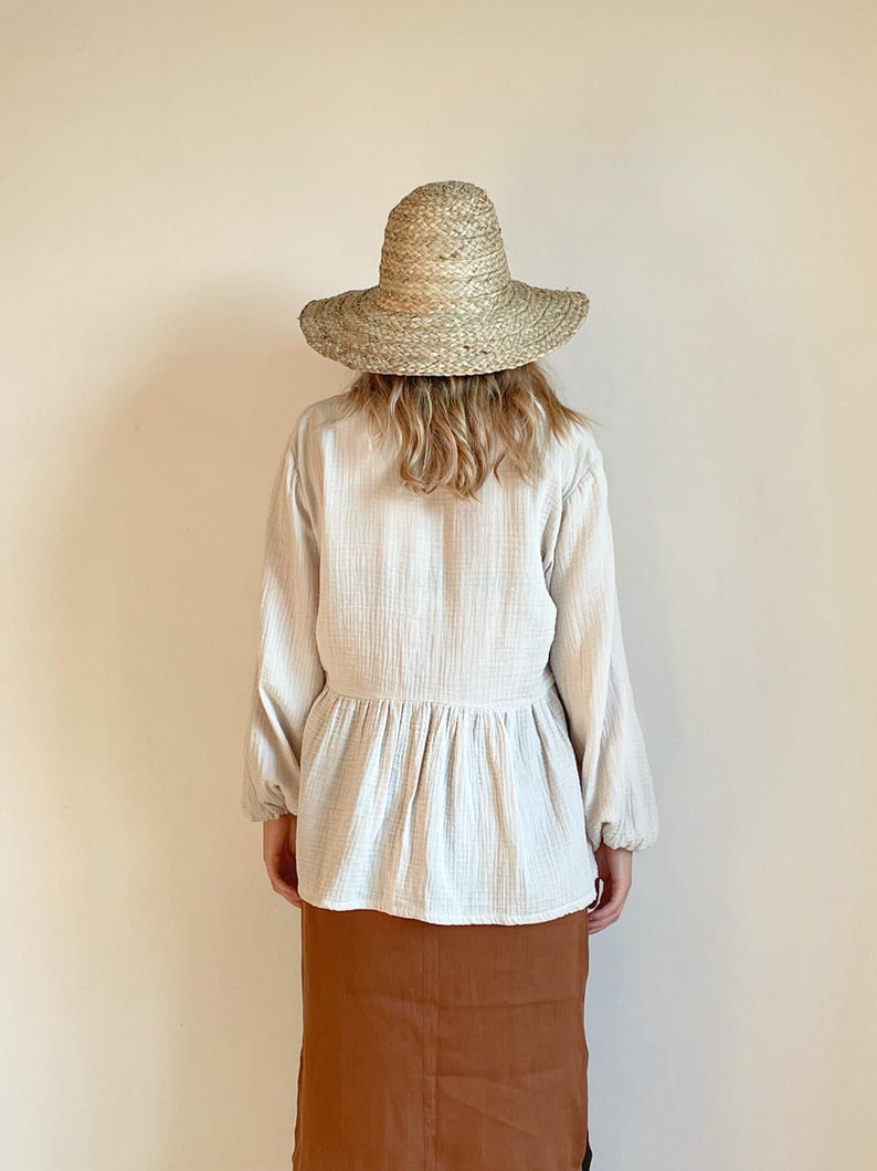 May include: A woman wearing a white, long-sleeved blouse with a gathered waist and a brown skirt. She is wearing a wide-brimmed straw hat.
