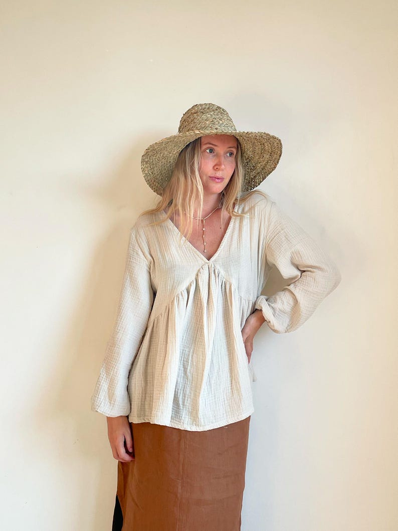 May include: A woman wearing a white, long-sleeved, loose-fitting blouse with a v-neck and a brown skirt. She is wearing a straw hat with a wide brim.