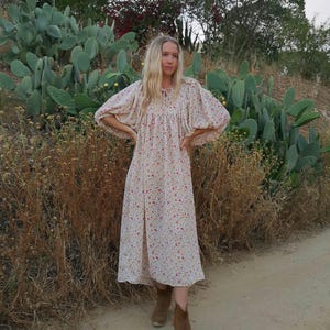 May include: A woman wearing a white floral midi dress with puffed sleeves and brown ankle boots. She is standing in a field with cacti.