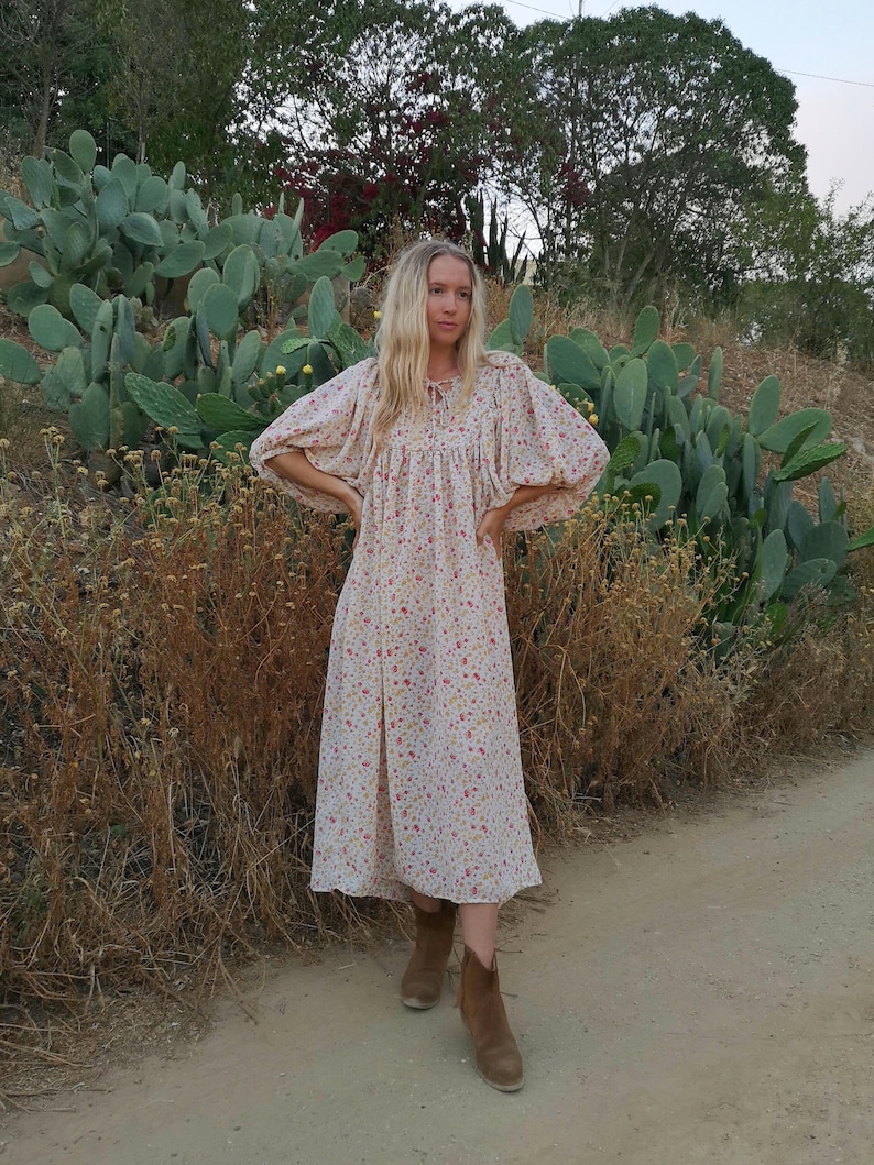 May include: A woman wearing a white floral midi dress with puffed sleeves and brown ankle boots. She is standing in a field with cacti.