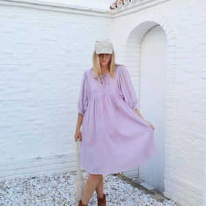 May include: A woman wearing a lavender dress with a white cap and brown ankle boots. She is standing in front of a white brick wall with a white door.