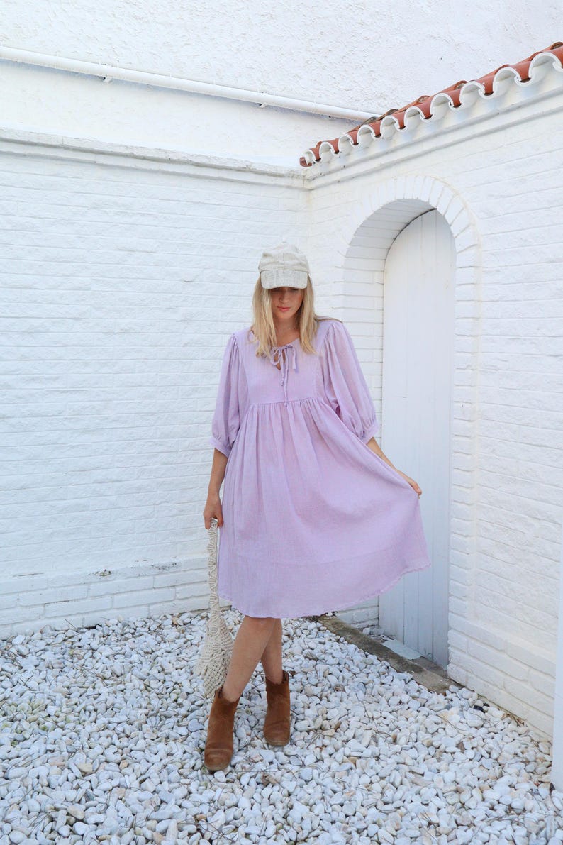 May include: A woman wearing a lavender dress with a white cap and brown ankle boots. She is standing in front of a white brick wall with a white door.