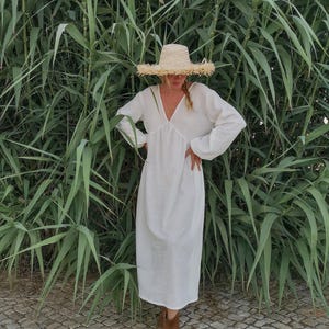 May include: A white maxi dress with long sleeves and a V-neckline. The dress is worn by a person wearing a straw hat and brown ankle boots. The person is standing in front of a wall of green plants.
