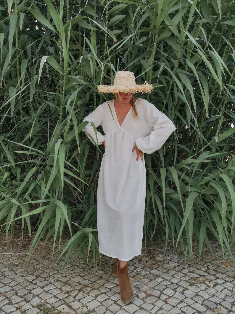 May include: A white maxi dress with long sleeves and a V-neckline. The dress is worn by a person wearing a straw hat and brown ankle boots. The person is standing in front of a wall of green plants.