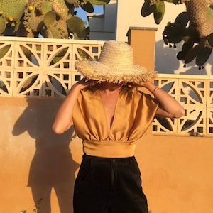 Pu&ograve; includere: Una donna indossa una blusa di raso dorata con scollo a V profondo e pantaloncini di velluto a coste neri. Indossa un grande cappello di paglia con una bordatura a frangia.