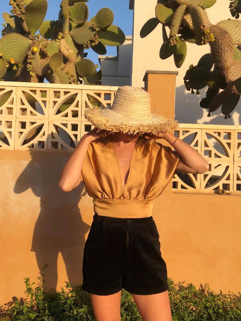 Pu&ograve; includere: Una donna indossa una blusa di raso dorata con scollo a V profondo e pantaloncini di velluto a coste neri. Indossa un grande cappello di paglia con una bordatura a frangia.
