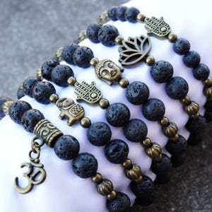 May include: Several black lava stone bead bracelets with antique gold-colored charms. Charms include an Om symbol, an elephant, a Buddha head, a lotus flower, and a Hamsa hand. The bracelets are displayed on a white surface.