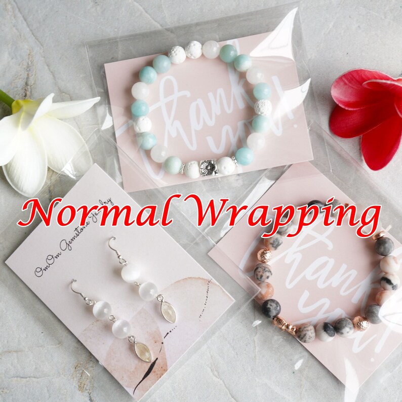 Op de afbeelding: Een close-up van drie sieraden verpakt in doorzichtige plastic zakken. De items omvatten een lichtblauwe en witte kralen armband, een paar witte stenen oorbellen en een roze en witte kralen armband. De zakken liggen op een wit oppervlak met een roze kaart waarop 'Bedankt' staat.