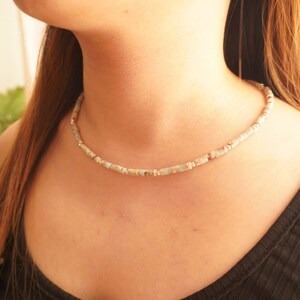 May include: A delicate necklace made of small, light blue and white patterned beads. The necklace is worn by a person with a light complexion.