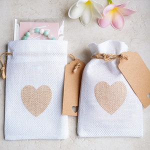 Op de afbeelding: Twee witte jute cadeauzakken met bruine papieren etiketten en een jute hartontwerp. Elke zak heeft een bruine touwband.