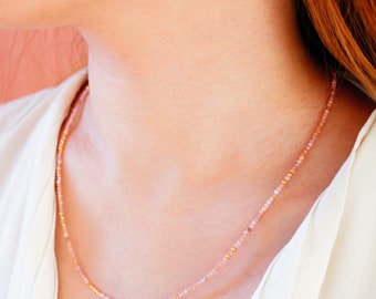 May include: A delicate necklace made of small pink and gold beads. The necklace is worn by a person with a white shirt.