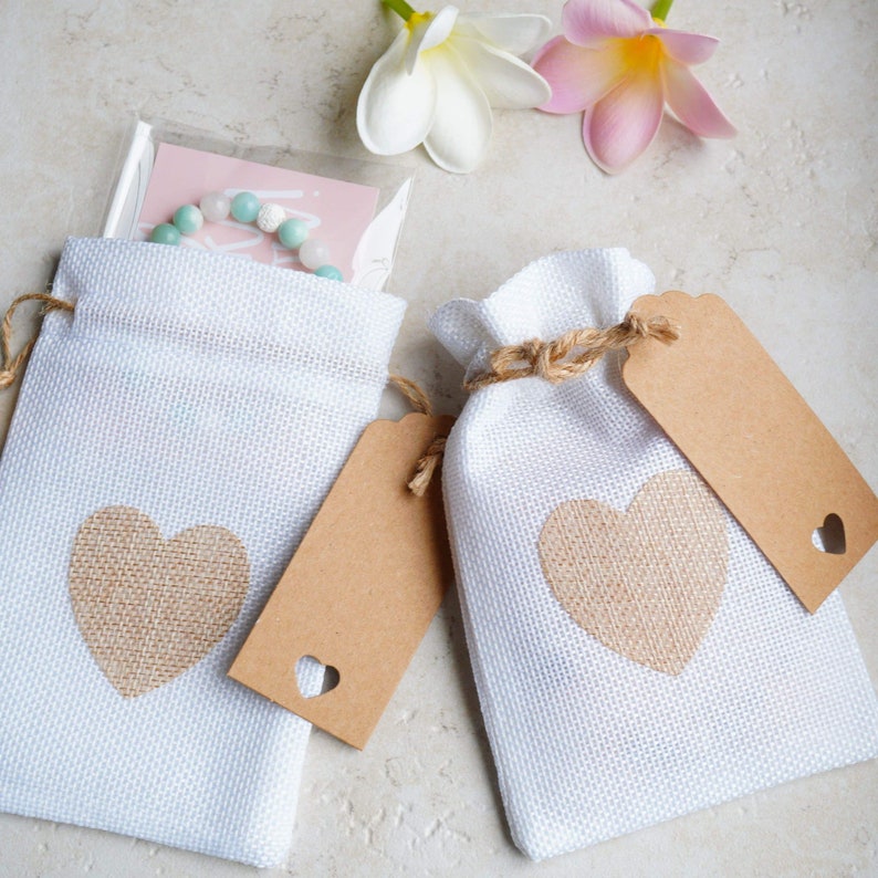 Op de afbeelding: Twee witte stoffen cadeauzakjes met bruine juteharten en bruine papieren kaartjes. De zakjes zijn vastgebonden met touw en hebben een rustieke, handgemaakte uitstraling.