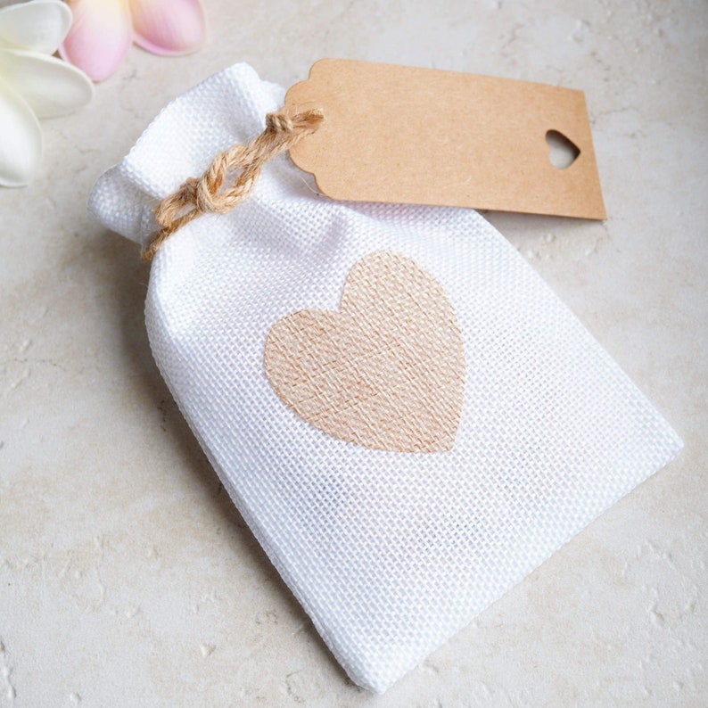 Op de afbeelding: Een witte stoffen cadeauzak met een bruin hartontwerp en een bruin papieren label met een hartvormige uitsparing. De zak is vastgebonden met touw.
