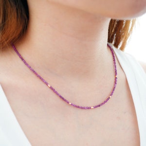 Peut inclure: Un collier délicat composé de petites perles de pierres précieuses violettes et d'accents dorés. Le collier est porté par une personne portant une chemise blanche.