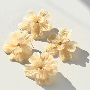 Könnte beinhalten: Vier zarte, blumenförmige Ohrringe in einem blassen Cremeton mit goldenen Akzenten. Die Ohrringe sind aus einem durchscheinenden Material gefertigt und haben eine leicht irisierende Oberfläche.