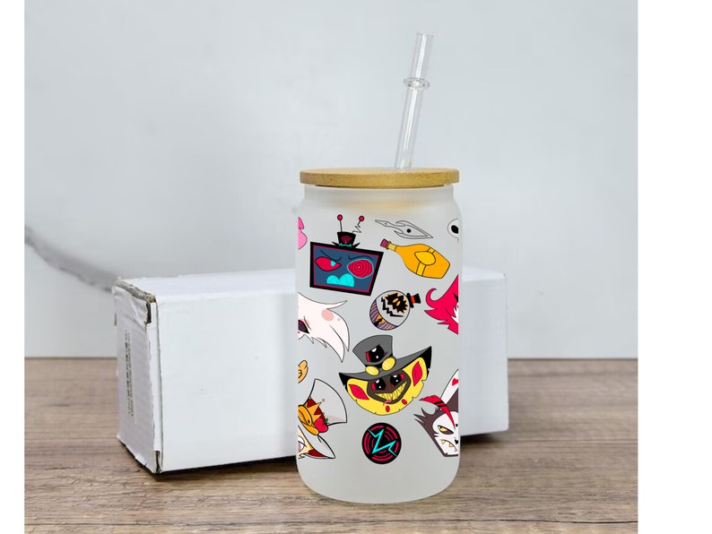 May include: A white frosted glass tumbler with a bamboo lid and a clear drinking straw. The tumbler is decorated with colorful cartoon characters and designs, including a red-eyed robot, a yellow character with a top hat, and a white character with wings.