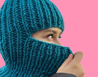 May include: A teal blue knitted balaclava with a face opening. The balaclava is made of thick yarn and has a ribbed texture.