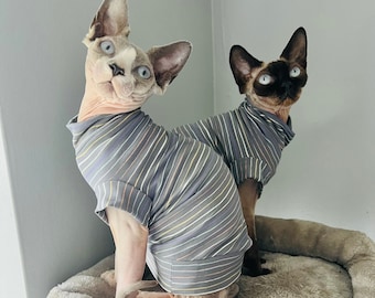 May include: Two hairless cats wearing matching striped shirts are sitting on a cat tree. The cat on the left is looking up and the cat on the right is looking to the side.