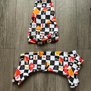 May include: Two-piece dog outfit with a black and white chequered pattern, adorned with red, orange, and yellow mushroom and eye motifs. The outfit is made of a soft, flexible material.