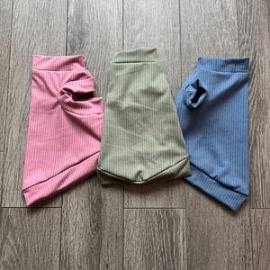Peut inclure: Trois chemises pour animaux de compagnie en tissu côtelé rose, vert olive et bleu. Chaque chemise a un col roulé et une ouverture pour les manches. Les chemises sont présentées sur une surface en bois.