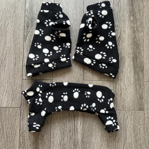 Könnte beinhalten: Schwarzer Tier-Pyjama mit weißen Pfotenabdrücken. Das Set besteht aus Ober- und Unterteil mit Öffnungen für Beine und Kopf. Der Stoff scheint weich und gemütlich zu sein, ideal zum Warmhalten von Haustieren.