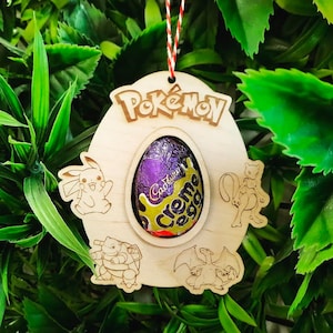 Może przedstawiać: Drewniany stojak na jajka Pokémon z jajkiem Cadbury Creme Egg w środku. Stojak ma wycięcie przedstawiające Pikachu, Charizard, Blastoise i Venusaur.