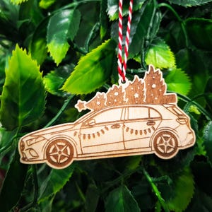 May include: Wooden Christmas ornament shaped like a car carrying Christmas trees on top. The car is decorated with a string of lights and has a red, white, and green string for hanging.