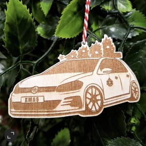 May include: Wooden car ornament with a Christmas tree design on top. The ornament is light brown with the word "KMA5" on the license plate. A red and white string is attached for hanging. The ornament is hanging in front of green leaves.