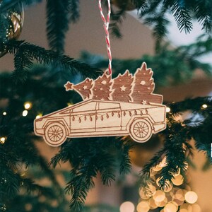 May include: A wooden Christmas ornament shaped like a pickup truck carrying a small Christmas tree on its bed. The truck is brown and has a white string hanging from the top.