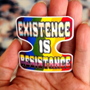 May include: A rainbow-colored sticker with the text "EXISTENCE IS RESISTANCE" in white letters. The sticker is designed to look like a puzzle piece.
