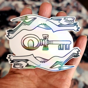May include: A holographic sticker featuring two white wolves running with a key in the middle. The key has a large circular head with a smaller keyhole and a long, thin shaft with a decorative handle.