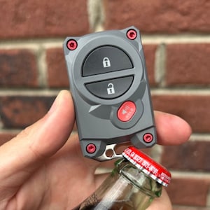 Könnte beinhalten: Ein grauer Autoschlüssel mit roten Akzenten und einem Flaschenöffner. Der Schlüssel hat zwei Verriegelungstasten und eine Paniktaste. Der Flaschenöffner wird verwendet, um eine Flasche Limonade zu öffnen.