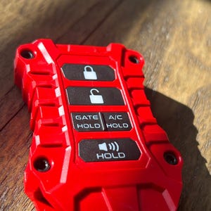 Może przedstawiać: Czerwony pilot zdalnego sterowania z czterema przyciskami. Przyciski są oznaczone jako "Gate Hold", "A/C Hold" i "Hold" z ikoną głośnika. Pilot ma teksturowaną powierzchnię i czarną plastikową obudowę.