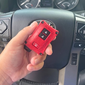 May include: A red car key fob with a textured surface and black buttons. The key fob has a lock and unlock button, and a "HOLD" button. The key fob is held in front of a car steering wheel.