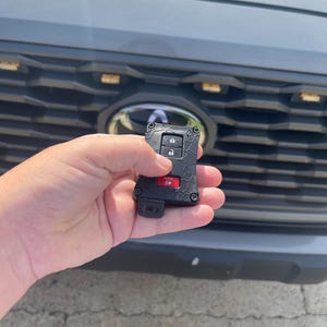 May include: Black car key fob with red and silver buttons. The key fob is held in front of a gray car's grill. The key fob has a textured design and the word "HOLD" is visible on a red button.