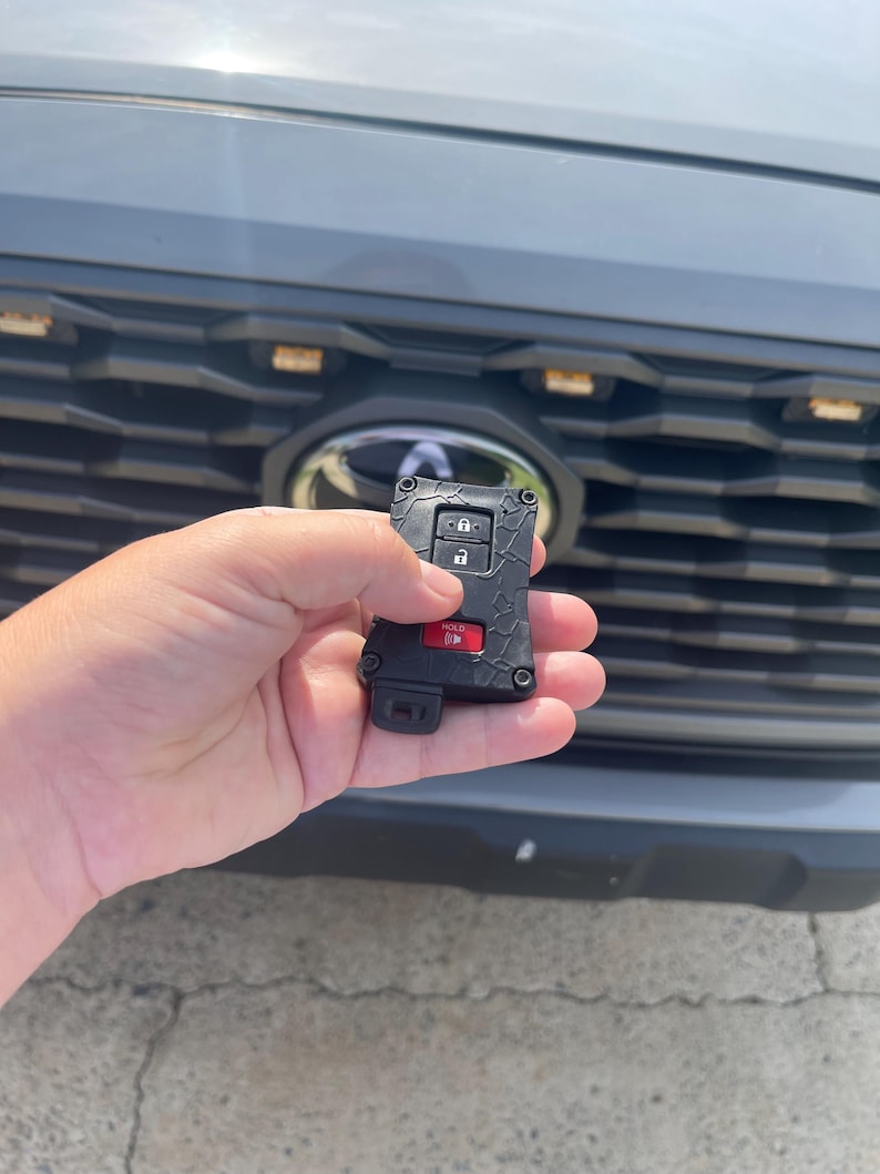 May include: Black car key fob with red and silver buttons. The key fob is held in front of a gray car's grill. The key fob has a textured design and the word "HOLD" is visible on a red button.