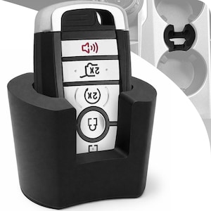 Puede incluir: Un llavero de coche negro con detalles plateados se sostiene en un soporte de espuma negro. El llavero tiene varios botones con iconos, incluyendo un altavoz, un candado y un símbolo de llave. El soporte está delante del interior de un coche.