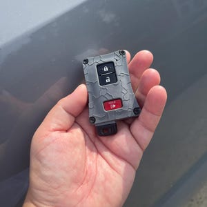 May include: A gray car key fob with a textured, cracked design. It features lock and unlock buttons, and a red "HOLD" button. The key fob is held in a person's hand.