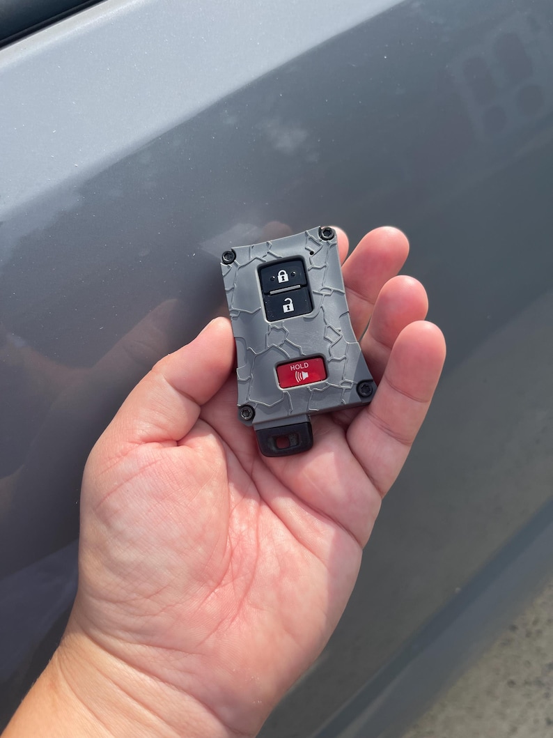 May include: A gray car key fob with a textured, cracked design. It features lock and unlock buttons, and a red "HOLD" button. The key fob is held in a person's hand.