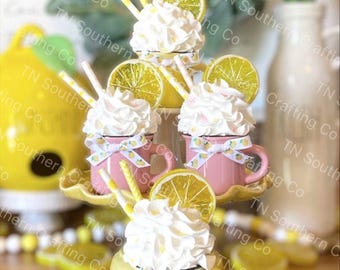 May include: Three miniature mugs with whipped cream and lemon slices on top. The mugs are decorated with yellow and pink ribbons and lemon print. The mugs are on a tiered stand.