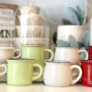 May include: A collection of eight mugs in various colors and styles, including white, red, green, and black. The mugs are arranged in a semi-circle on a table. Some of the mugs have a black rim and handle.