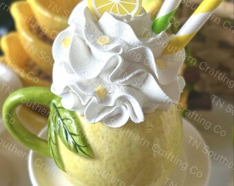 May include: A yellow ceramic mug shaped like a lemon with a white whipped cream topping and a lemon slice on top. The mug is sitting on a white saucer and has a green leaf handle. Two striped straws are sticking out of the whipped cream.