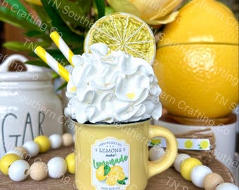 May include: Yellow ceramic mug with a white rim and a handle. The mug has a lemon-themed design with the text "Turn life's sour lemons into lemonade". The mug is topped with white whipped cream and a lemon slice.